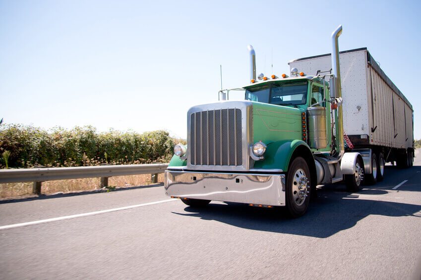 Classic American bonneted large green rig semi truck with high stylish chrome exhaust pipes transporting commercial cargo in bulk container trailer on the highway with trees behind the safety fence