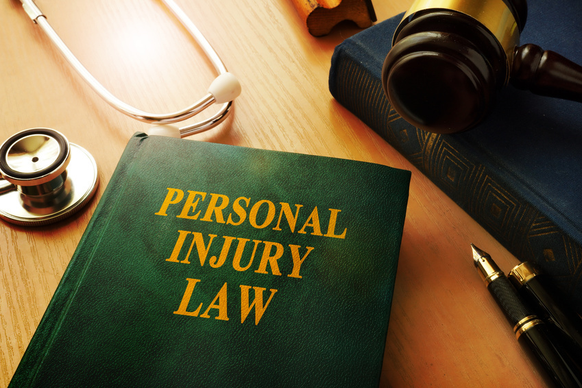 Green hard-bounded book with a title of "PERSONAL INJURY LAW" is placed on top of a wooden table along with a stethoscope, traditional stamp, black pen, gavel and a book