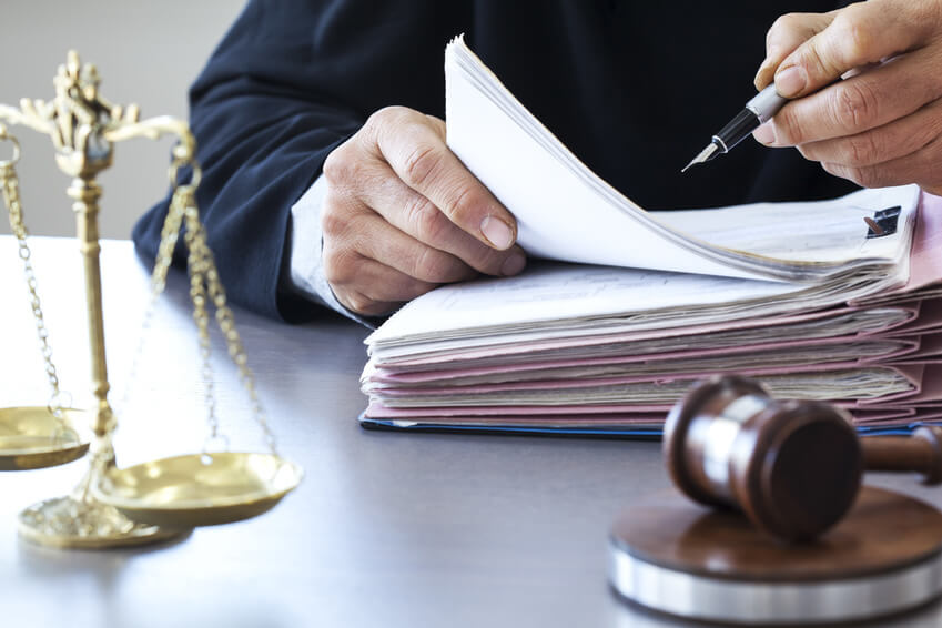 A man wearing a black robe like a best serious injury attorney dress is sitting and holding a fountain pen and writing on a thick pile of paperwork that is attached on a folder and is placed on top of a wooden table along with the block, gavel and golden scales of justice.